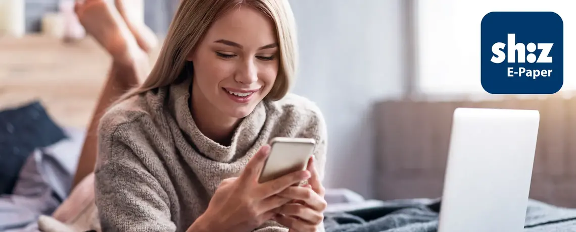 Junge Frau liegt auf einem Bett und liest auf ihrem Smartphone die digitale Zeitung in der App shz E-Paper
